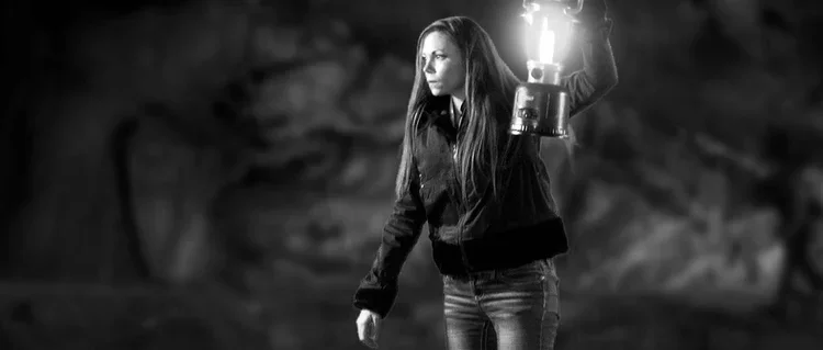 A woman with long hair holding a lantern in a dark, outdoor setting.