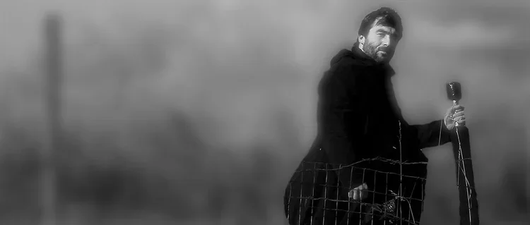 A man who is a LARPer standing outdoors with a foam sword, wearing a dark jacket, and a wire fence in front of him, in black and white.