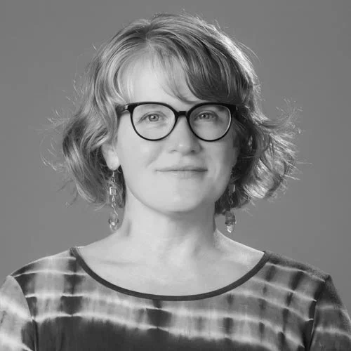 Black and white portrait of a woman with short, wavy hair, glasses, and earrings, wearing a patterned top, against a plain background.