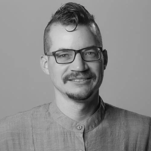 Black and white portrait of a man with glasses, short hair, and a goatee, wearing a high-collared shirt, smiling at the camera.