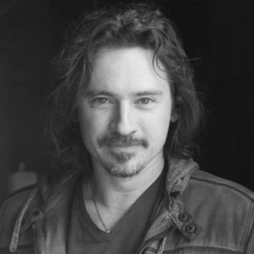 Black and white portrait of a man with long, wavy hair, a mustache, and a beard, wearing a jacket over a T-shirt, looking at the camera with a slight smile.