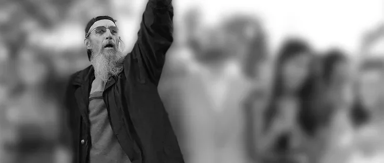 A man with a beard and sunglasses raising his arm, with a blurred outdoor crowd in the background.