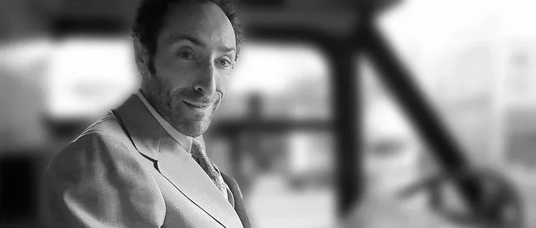 A man with dark curly hair and a beard, smiling, wearing a vest, sitting in a bus with a blurred background.
