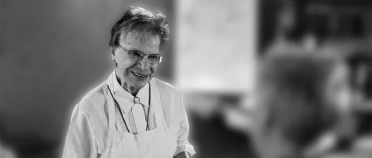 A happy elderly woman wearing glasses, a white apron, and a lab coat in a kitchen.