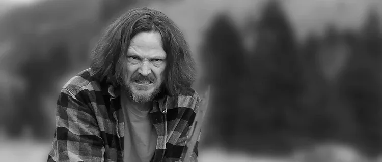 A man with long, messy hair and a beard, wearing a checkered shirt, making an angry or intense facial expression outdoors