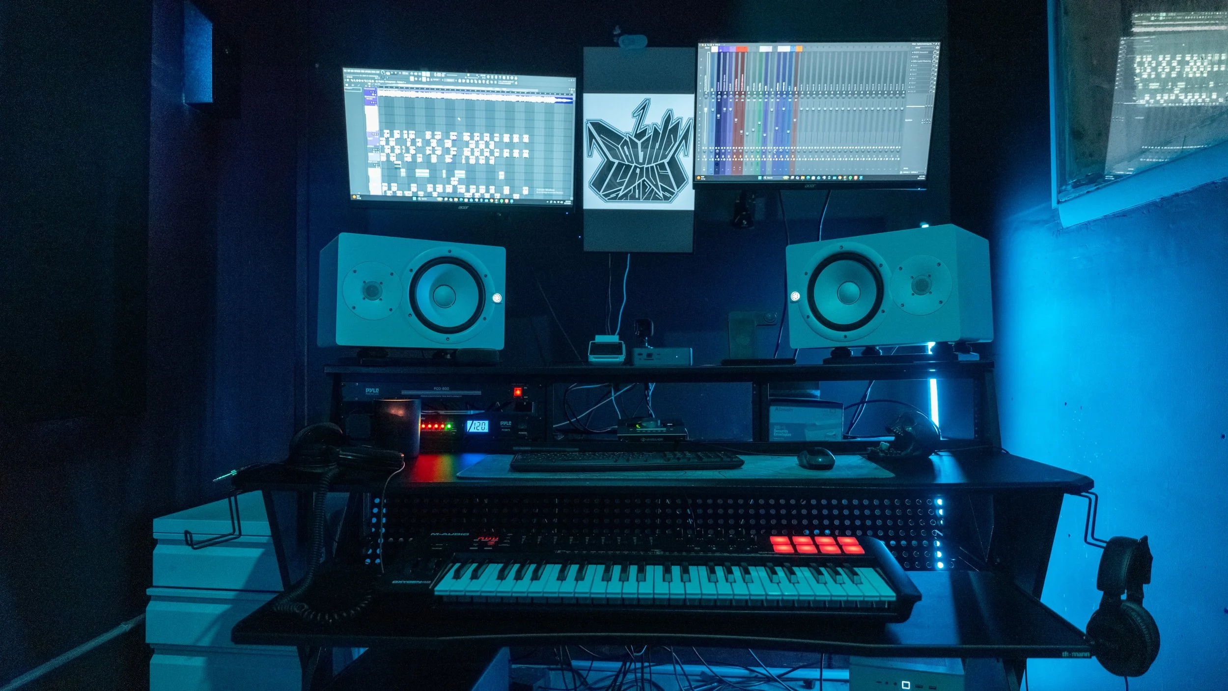 A music production setup with two monitors displaying digital audio workstations, a keyboard, studio speakers, a MIDI controller, and various electronic equipment illuminated by blue lighting.