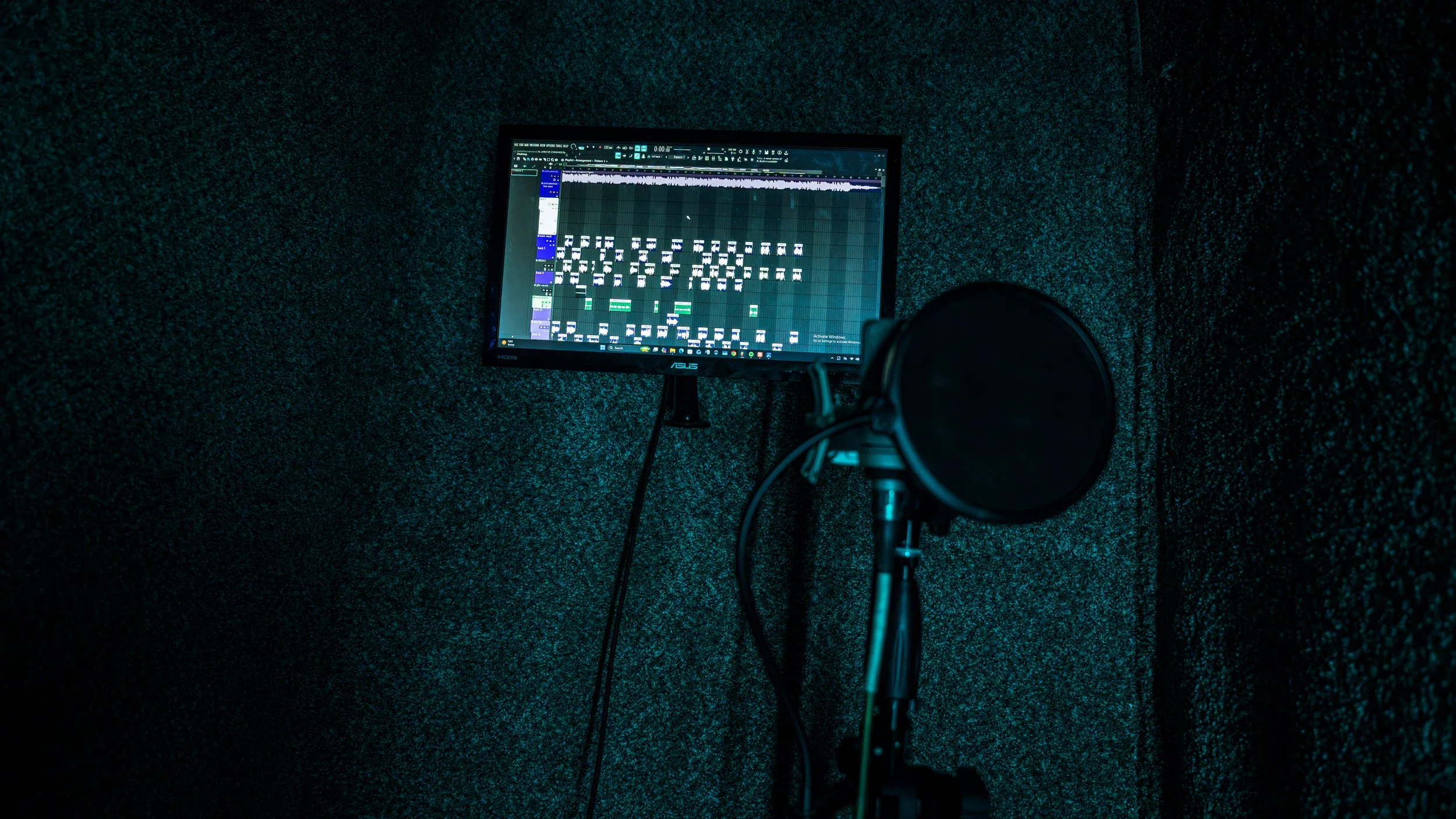 Music production studio with a computer monitor displaying audio editing software and a microphone in a soundproof room.