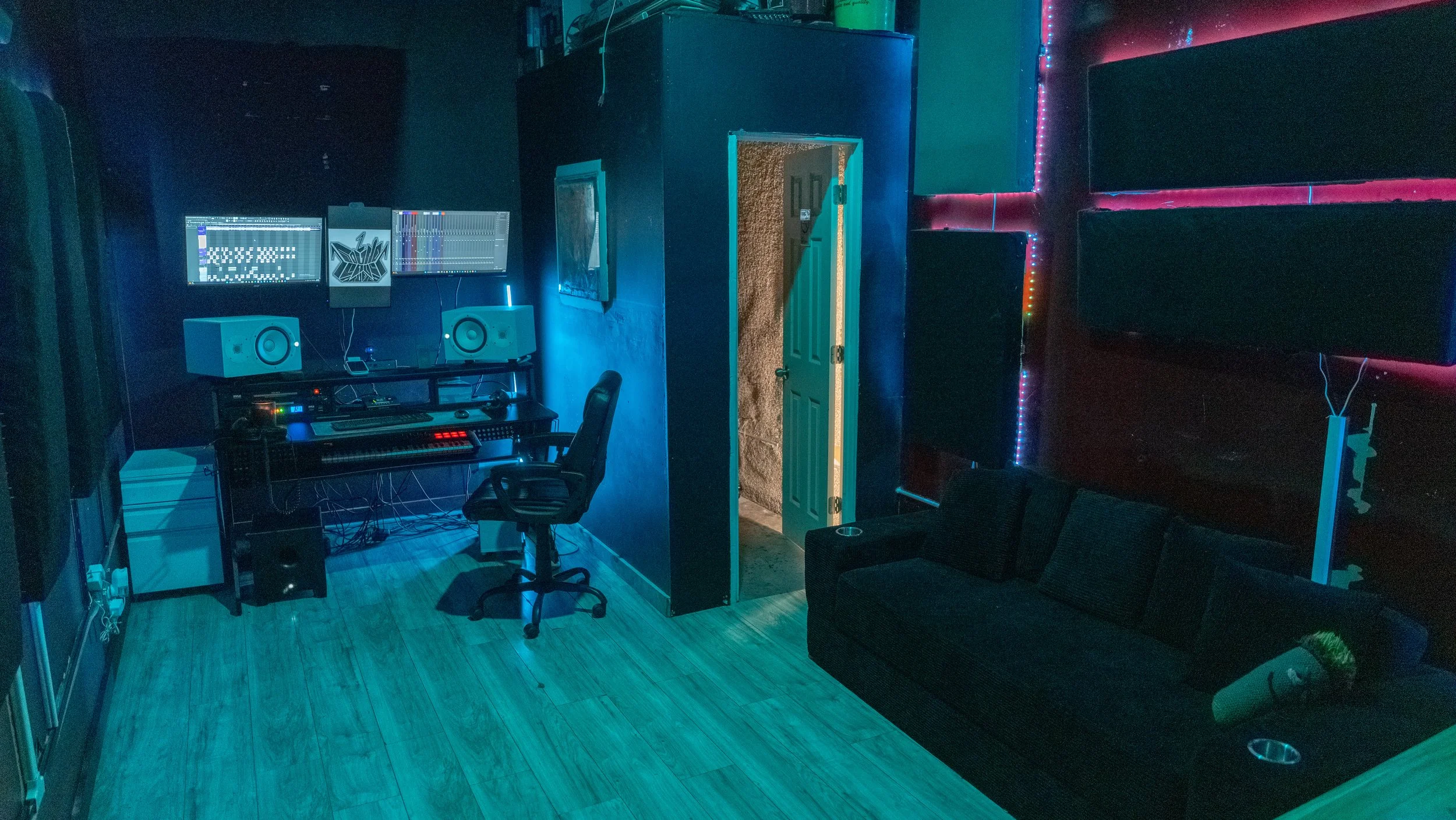 A music studio with two computer monitors and speakers on a desk, illuminated by blue and purple LED lights, with a black sofa on the right side.