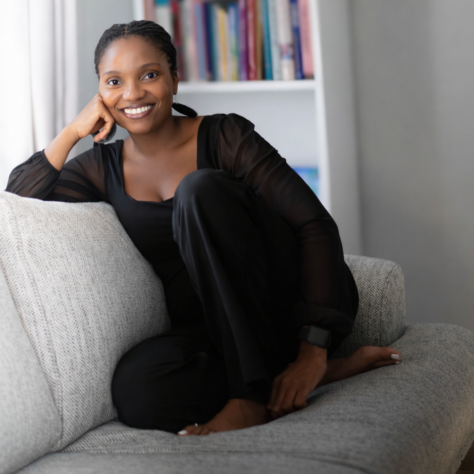 A woman with dark hair and a bright smile sitting on a light gray sofa in a cozy room with a bookshelf in the background.
