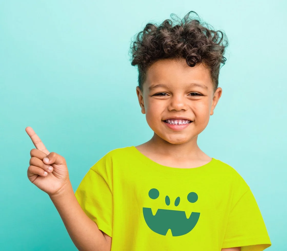 A smiling young boy with curly hair wearing a bright yellow T-shirt with a cartoon monster face, pointing upwards with his index finger against a light blue background.