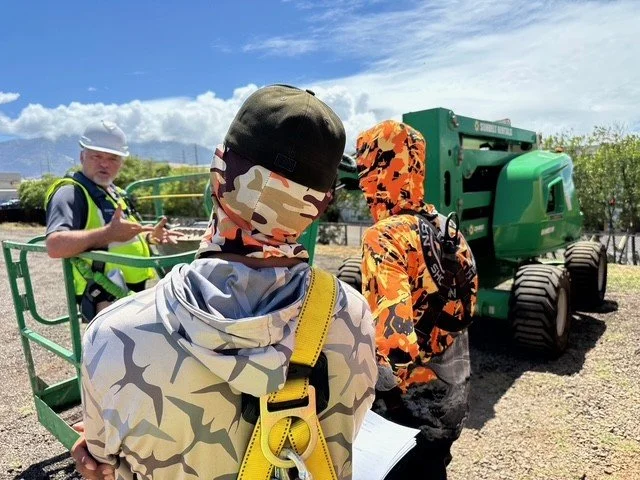 Two young boys dressed in camouflage hoodies and hats with safety harnesses are observing a green construction vehicle while a man explains something in the background on a sunny day.