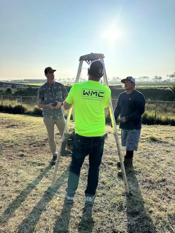 Three men standing outdoors near a fining instrument set up on a tripod, with a chain-link fence, grassy field, and trees in the background on a sunny day.