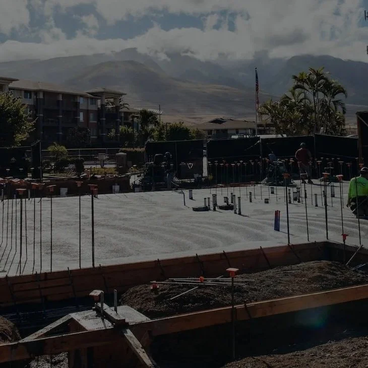 WMC Construction site with rebar and concrete foundation, buildings and trees in the background, mountains and cloudy sky in the distance.
