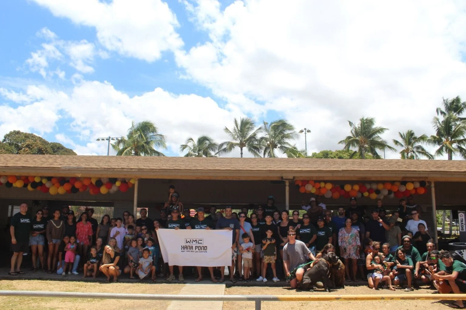 Group of people gathered outdoors under a pavilion decorated with orange, red, and yellow balloons, with palm trees and a partly cloudy sky in the background.