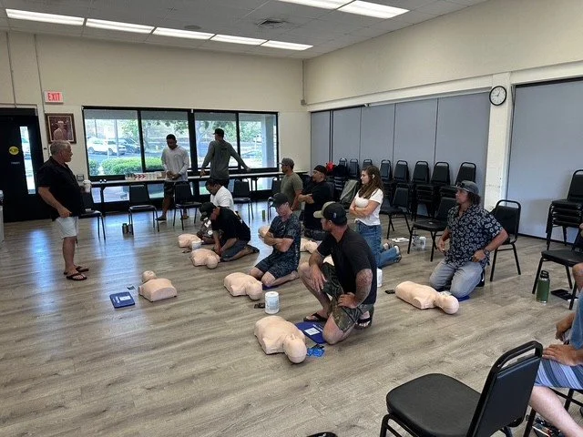 Group of people participating in a CPR training class with mannequins in a room with large windows and stacked chairs.