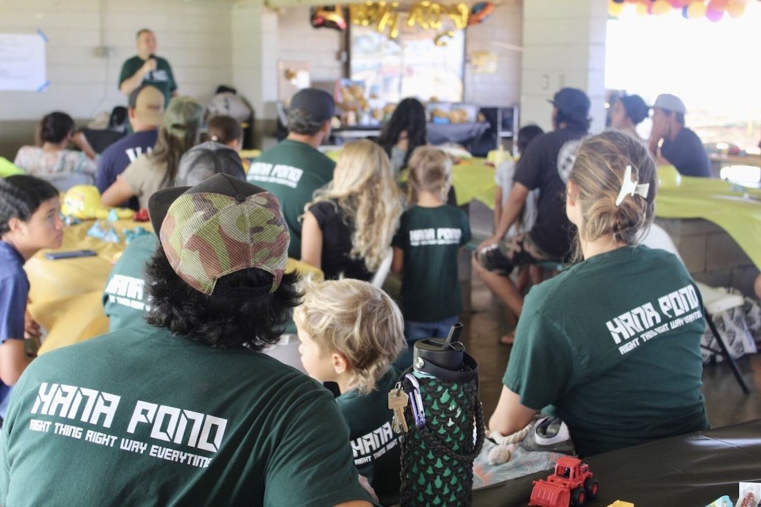 Group of people, including children and adults, seated at tables listening to a speaker in a room decorated for a celebration.