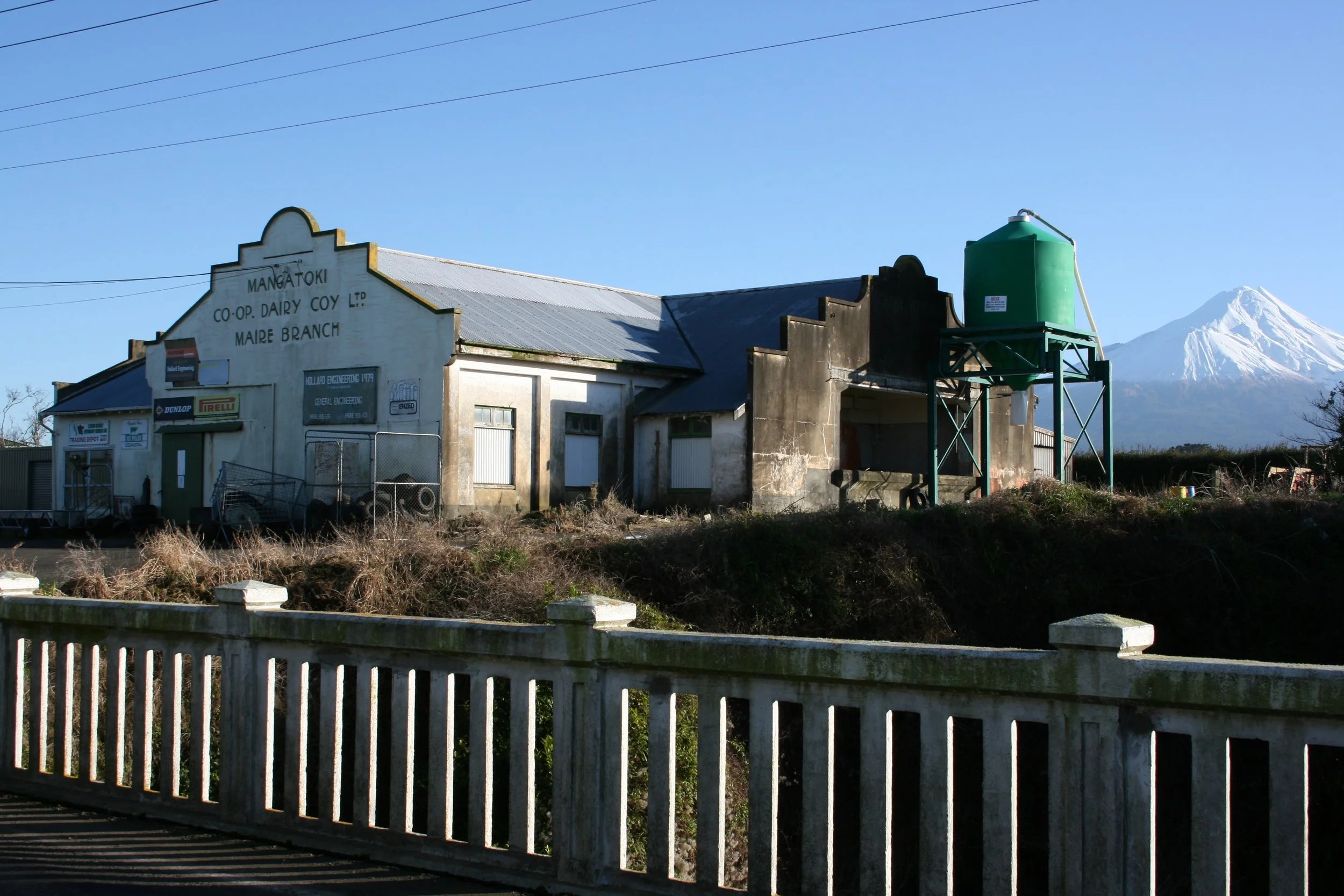 Building with a sign that reads 'MANGATOKI CO-OP DAIRY COY LTD MAIRE BRANCH' and a green water tank on a metal stand, with a mountain in the background.