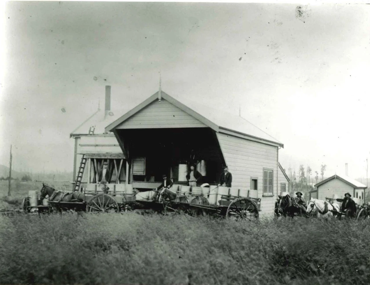 Black and white photograph of a barn with a group of people and horses in front of it, some standing on wagons.