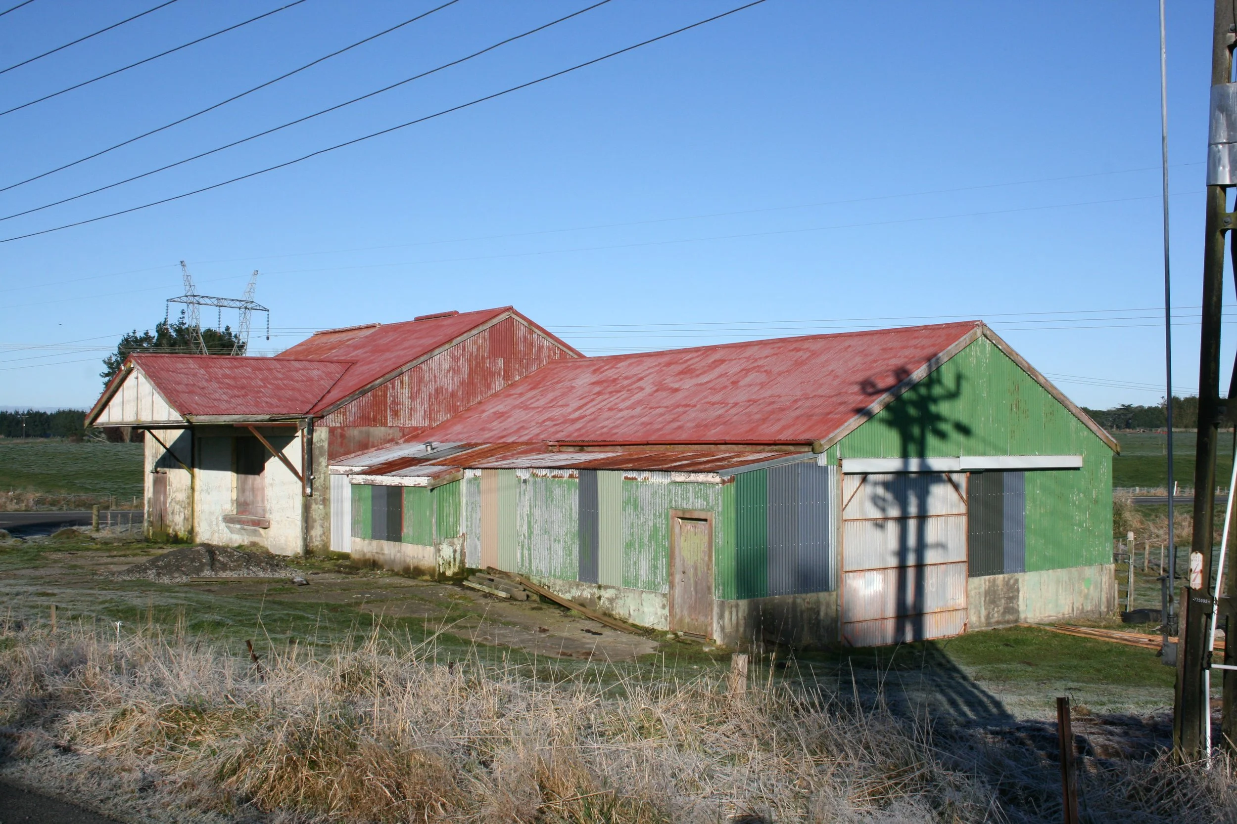 An old, rusty barn with a red and green metal roof in a rural area under a clear blue sky. Power lines run across the top of the image, and there is a shadow of a utility pole on the barn. The surrounding area has dry grass and dirt.