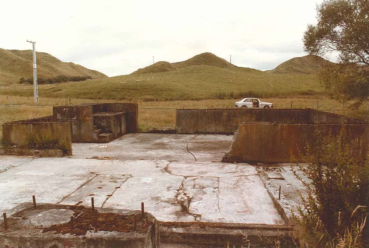 Ruined concrete foundation with rusted barriers, a white car in the background, grassy hills, a lone tree, and cloudy sky.