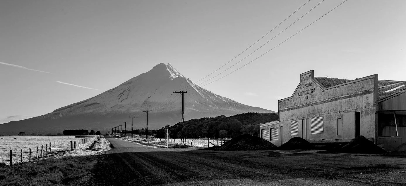 Black and white photo of Mount Taranaki in New Zealand with a road, utility poles, and an old building on the right.