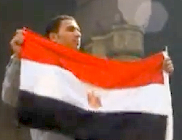 Young child wearing a hat and shirt with Arabic script, being carried on an adult's shoulders during a celebration with Egyptian flags and other children around.
