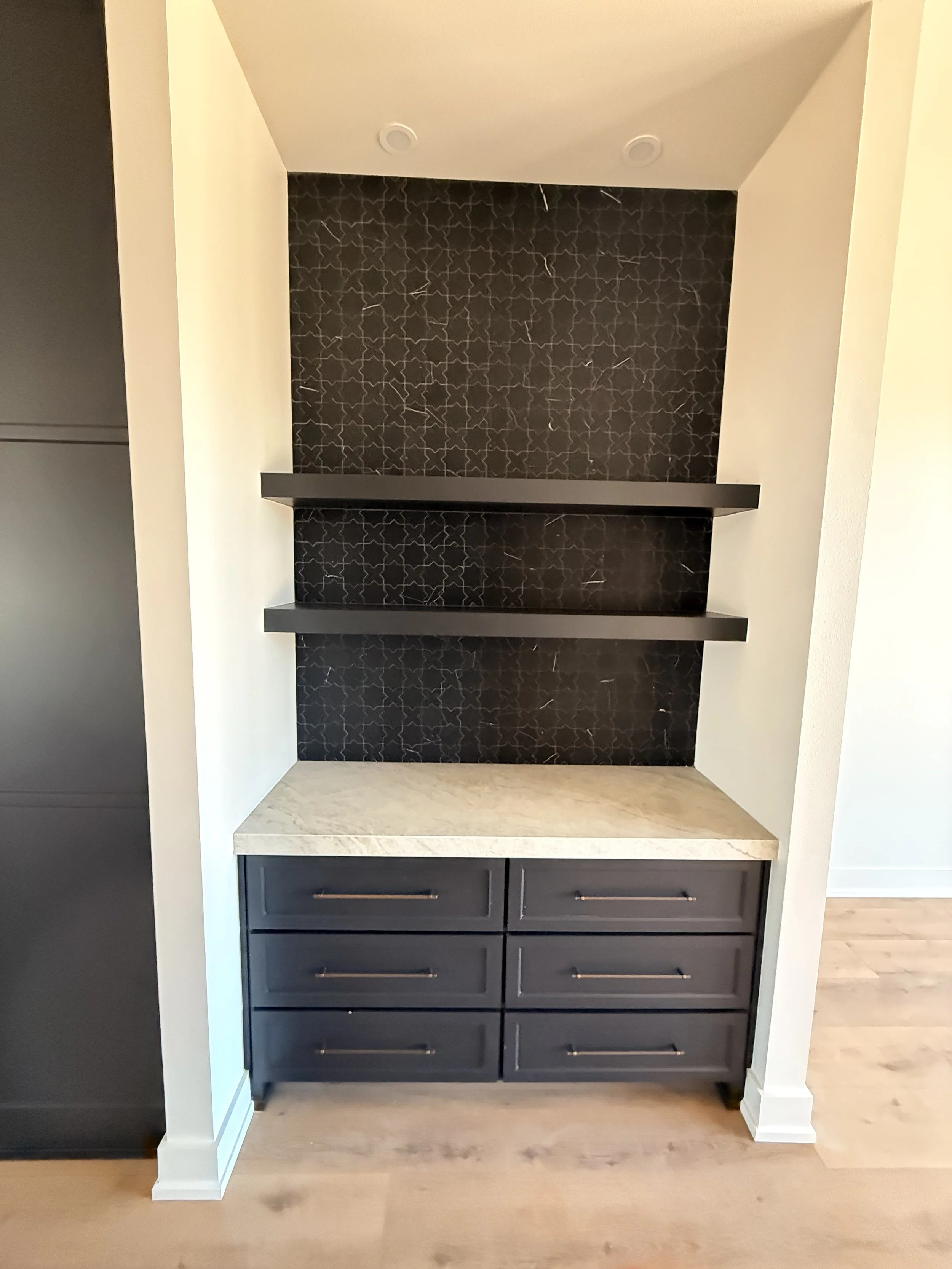 Built-in kitchen or bar area with dark gray drawers, a light-colored marble countertop, and two black floating shelves against a black patterned backsplash.