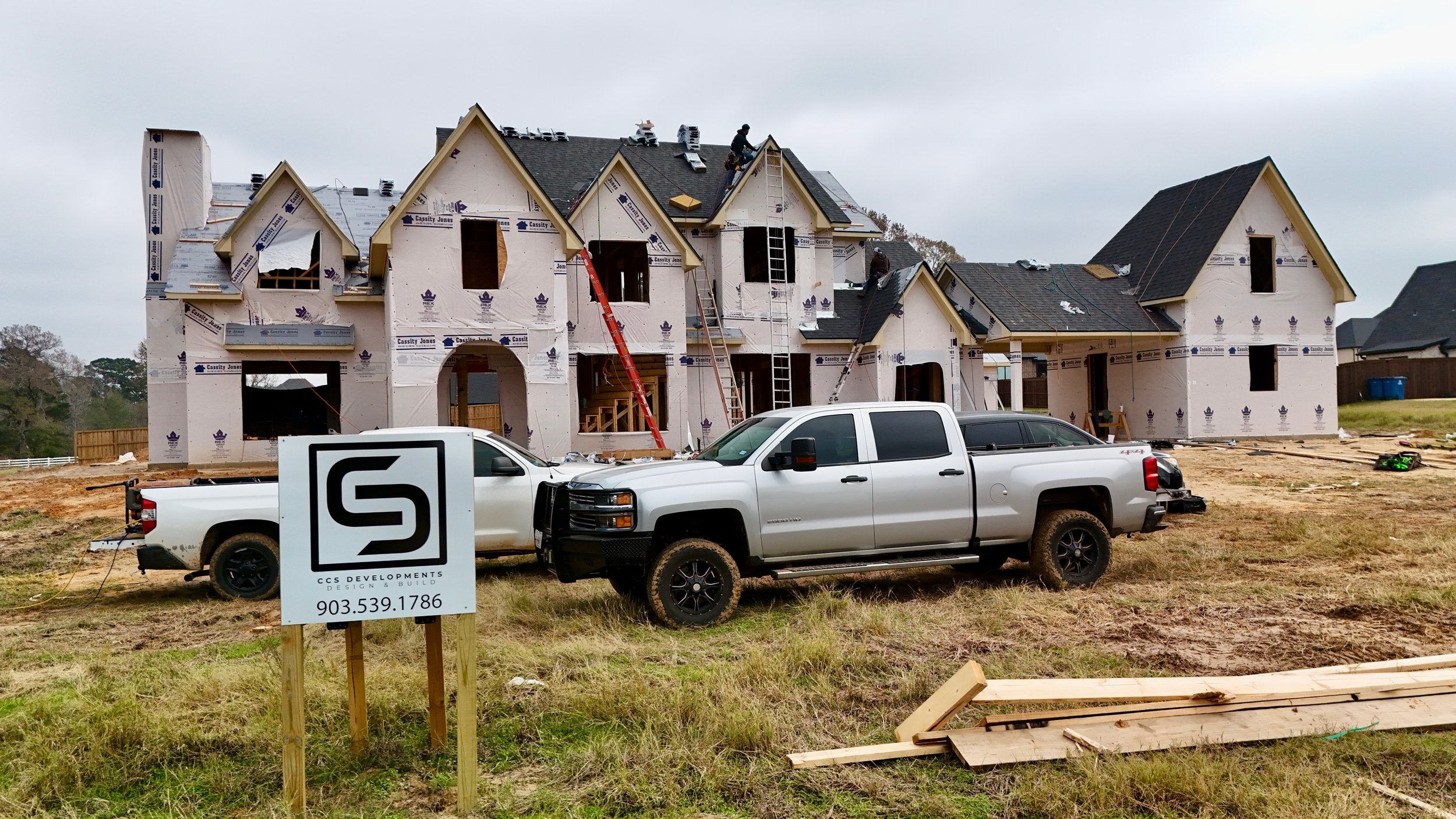 Construction site of a large house with workers on the roof, surrounded by vehicles, with wooden framing and building materials visible.