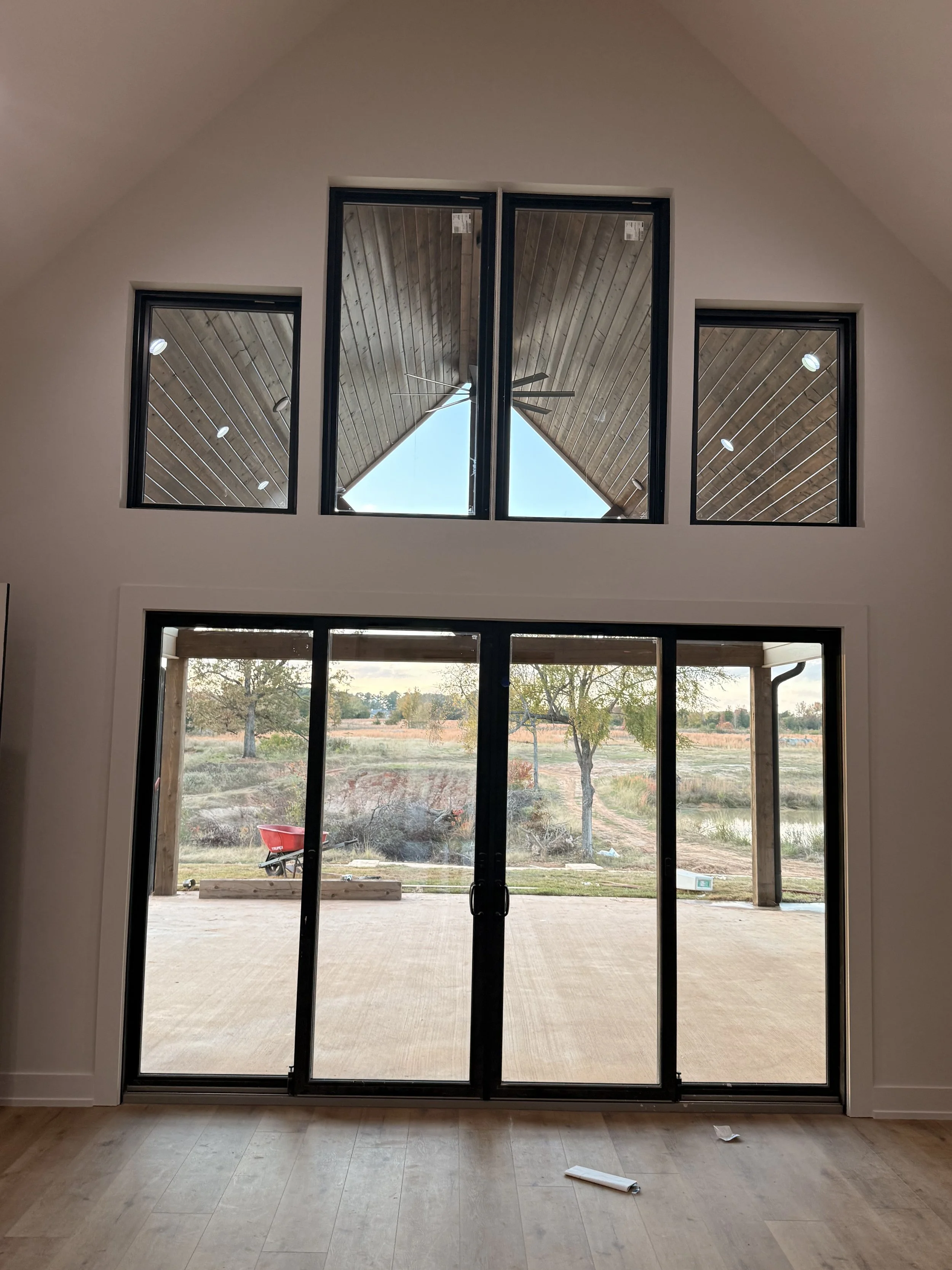 Interior view of a room with large sliding glass doors opening to an outdoor patio and landscape, and two tall windows above the doors revealing a wooden ceiling and outdoor scenery including trees and a wheelbarrow.
