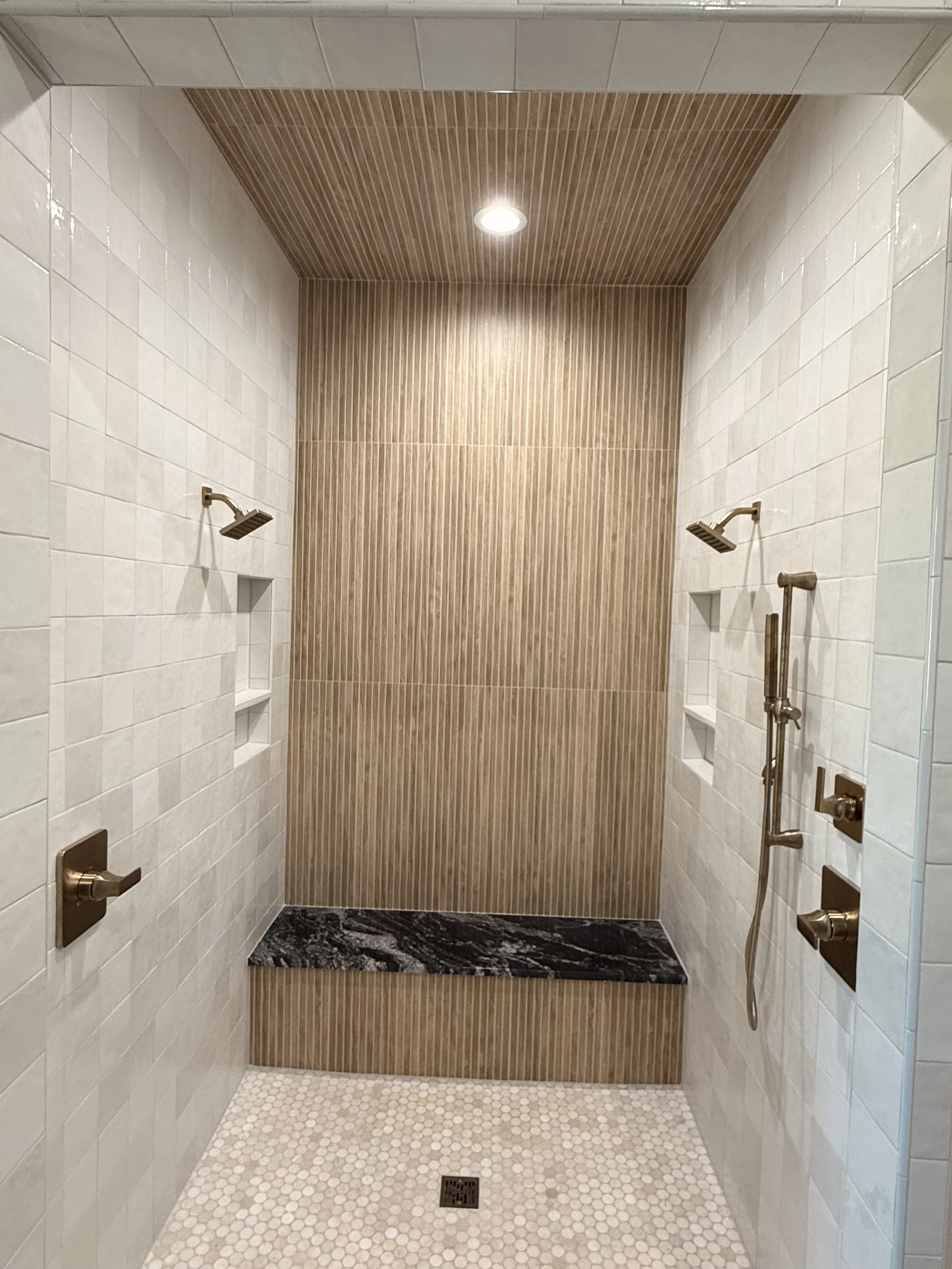 A modern walk-in shower with white tile on the sides, a brown wood-textured accent wall and ceiling, a built-in black marble bench, and gold fixtures including two showerheads, controls, and a vertical grab bar.