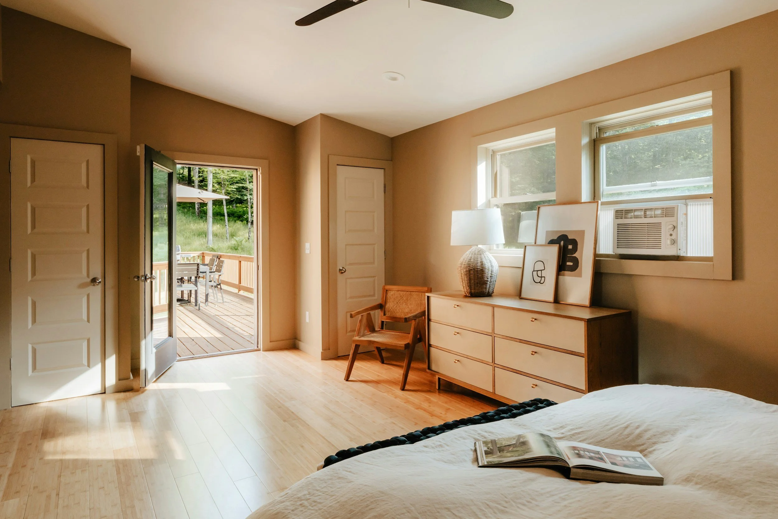 A bedroom with natural light, a wooden dresser with framed artwork and a lamp, a chair, and a door leading to a wooden deck with outdoor furniture, and windows with an air conditioning unit.