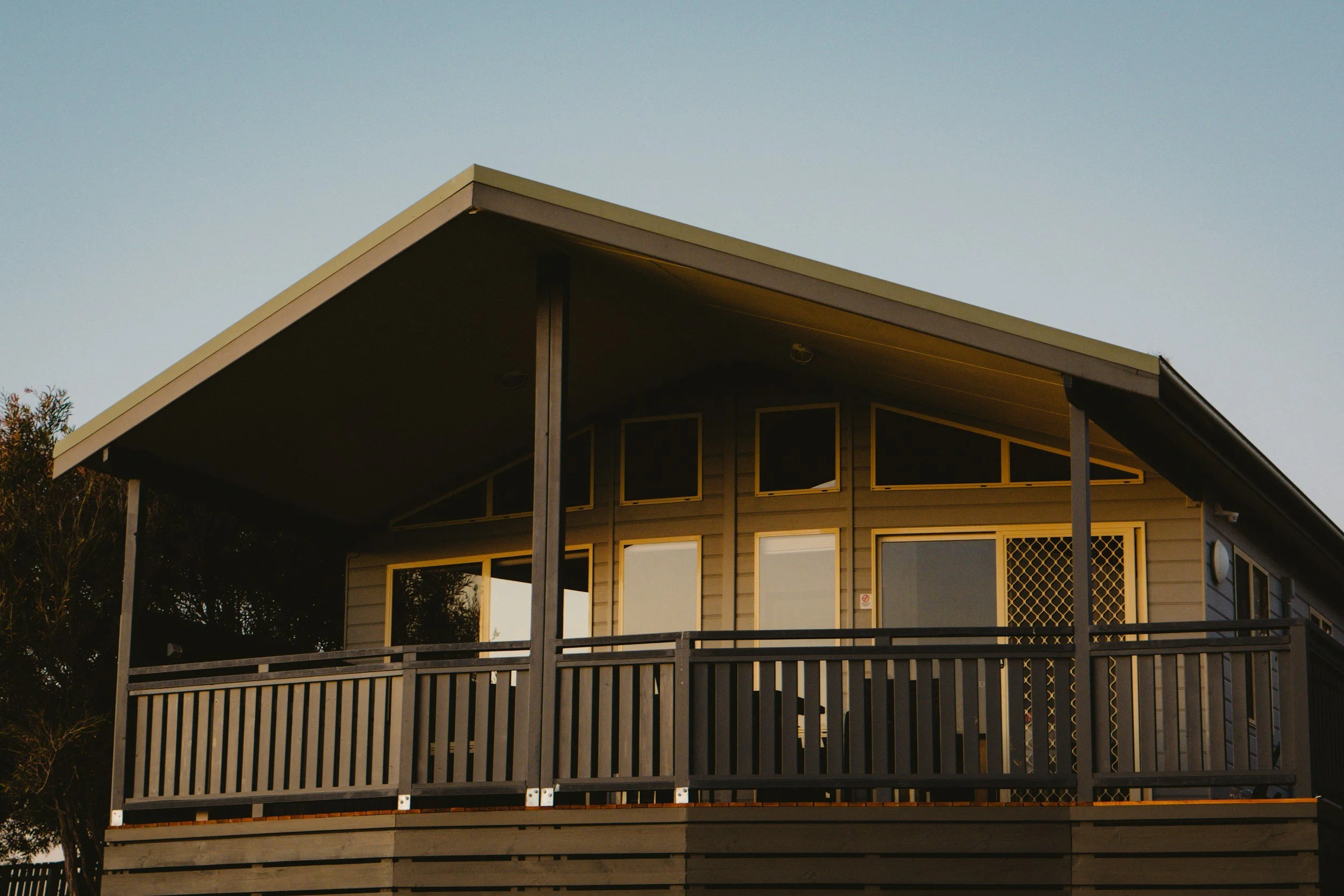 A modern house with a sloped roof and a front porch, featuring several large windows and a railing, during sunset.