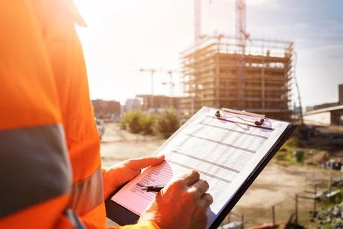 Construction worker in an orange safety vest reviewing documents or plans at a construction site with a building under construction and a crane in the background.