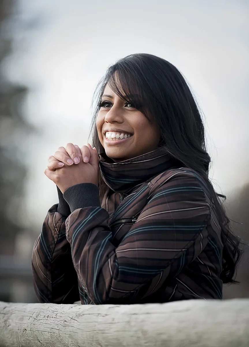 A young woman with long dark hair smiling and looking to the side outdoors, wearing a dark striped jacket, with hands clasped together resting on a wooden railing.