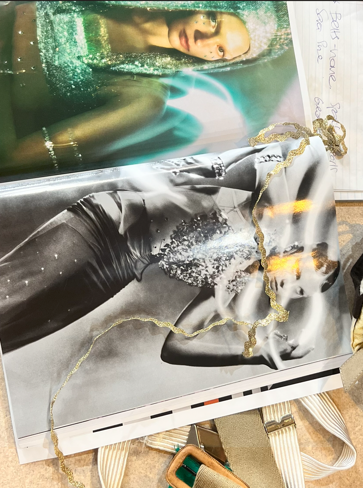 Photograph of two posters, one in color and one in black and white, featuring women. The color poster shows a woman with long blonde hair wearing a shiny green top, while the black and white poster shows a woman with long hair in a black dress with silver embellishments. Gold decorative string is draped across the posters. A notepad with handwritten notes is partially visible in the upper right corner.