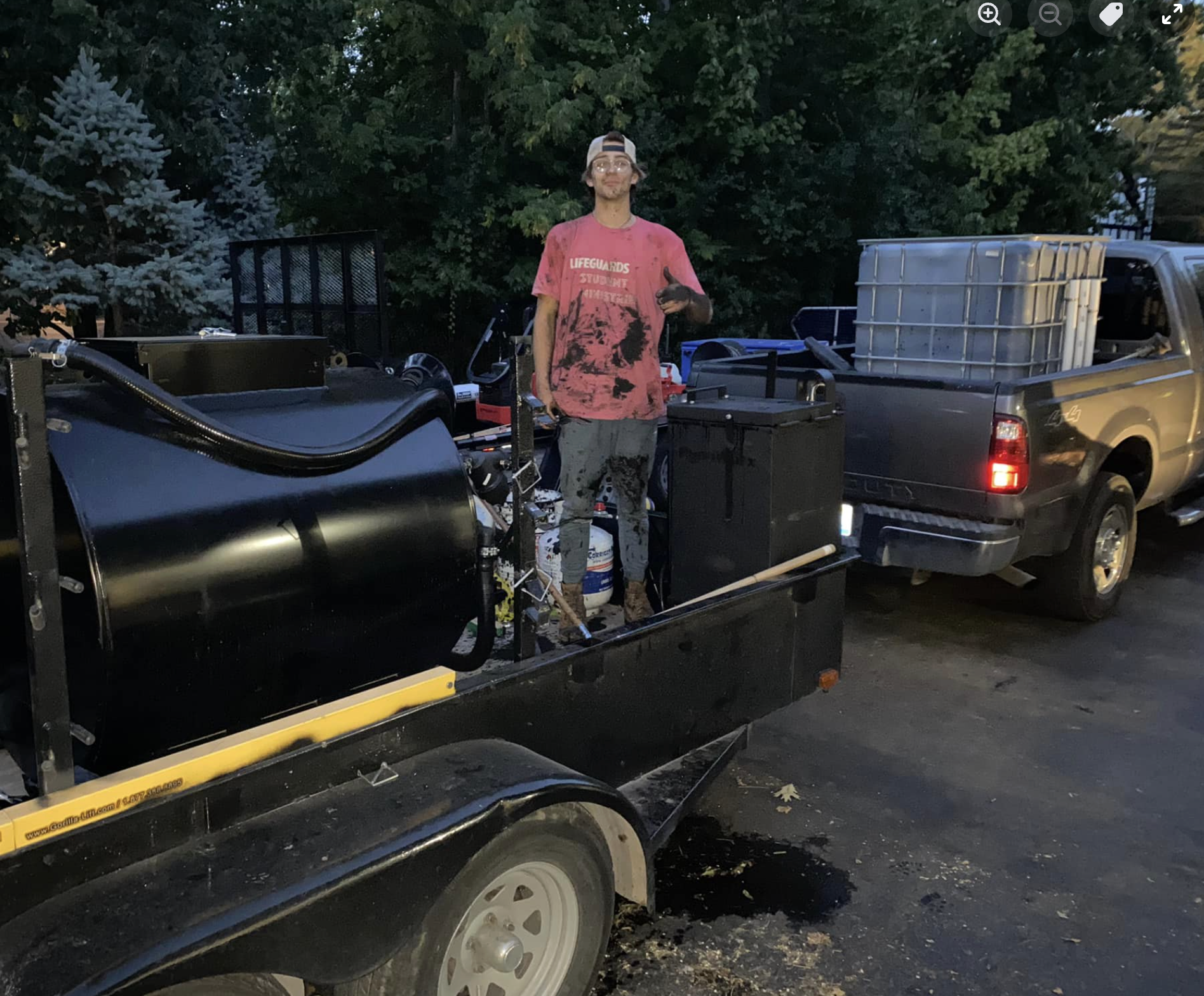 A young man standing on a trailer filled with equipment, including a large black tank and black boxes, parked beside a gray pickup truck with a large water tank in the truck bed, with trees in the background.