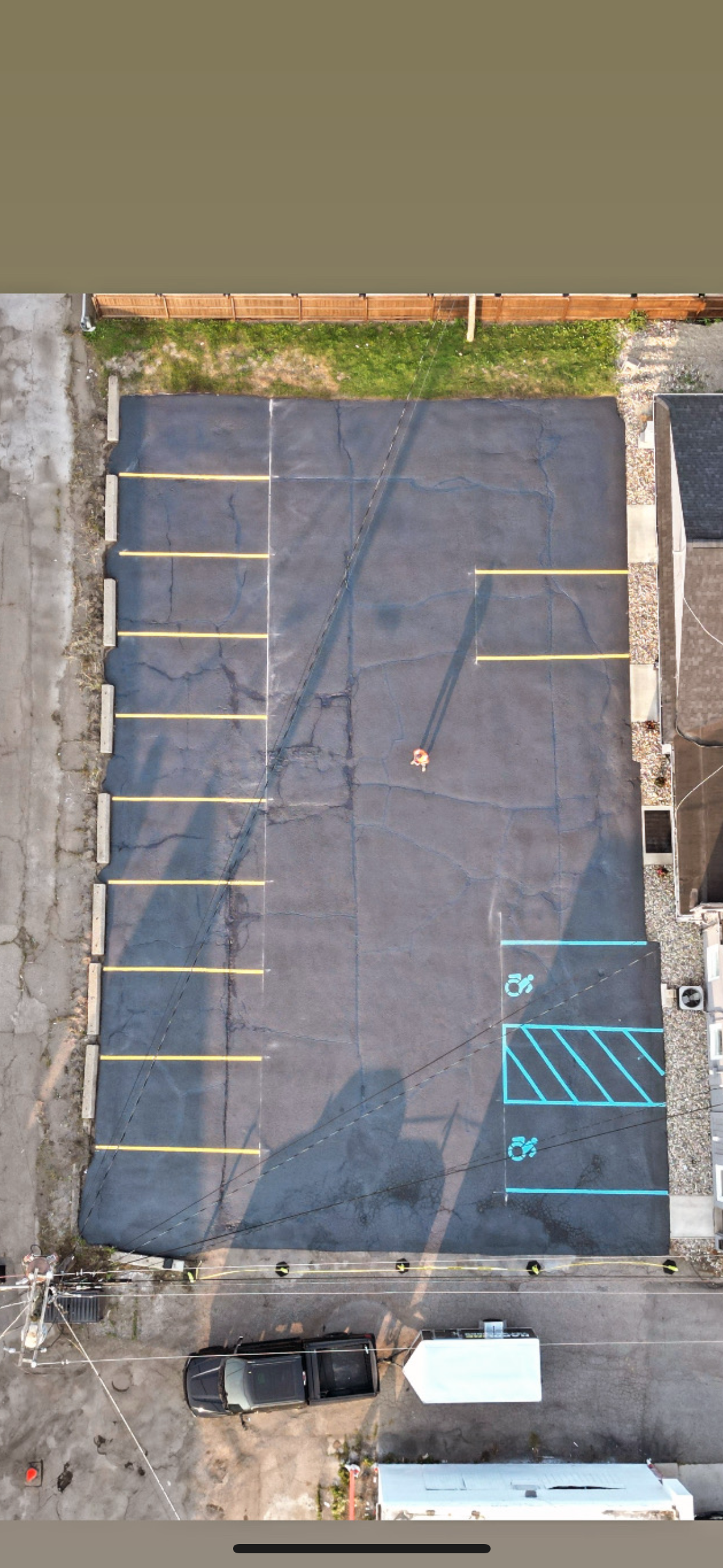 An empty parking lot with marked parking spaces, including two accessible spots in blue, surrounded by a wooden fence at the top, a building on the right, and a vehicle parked at the bottom.