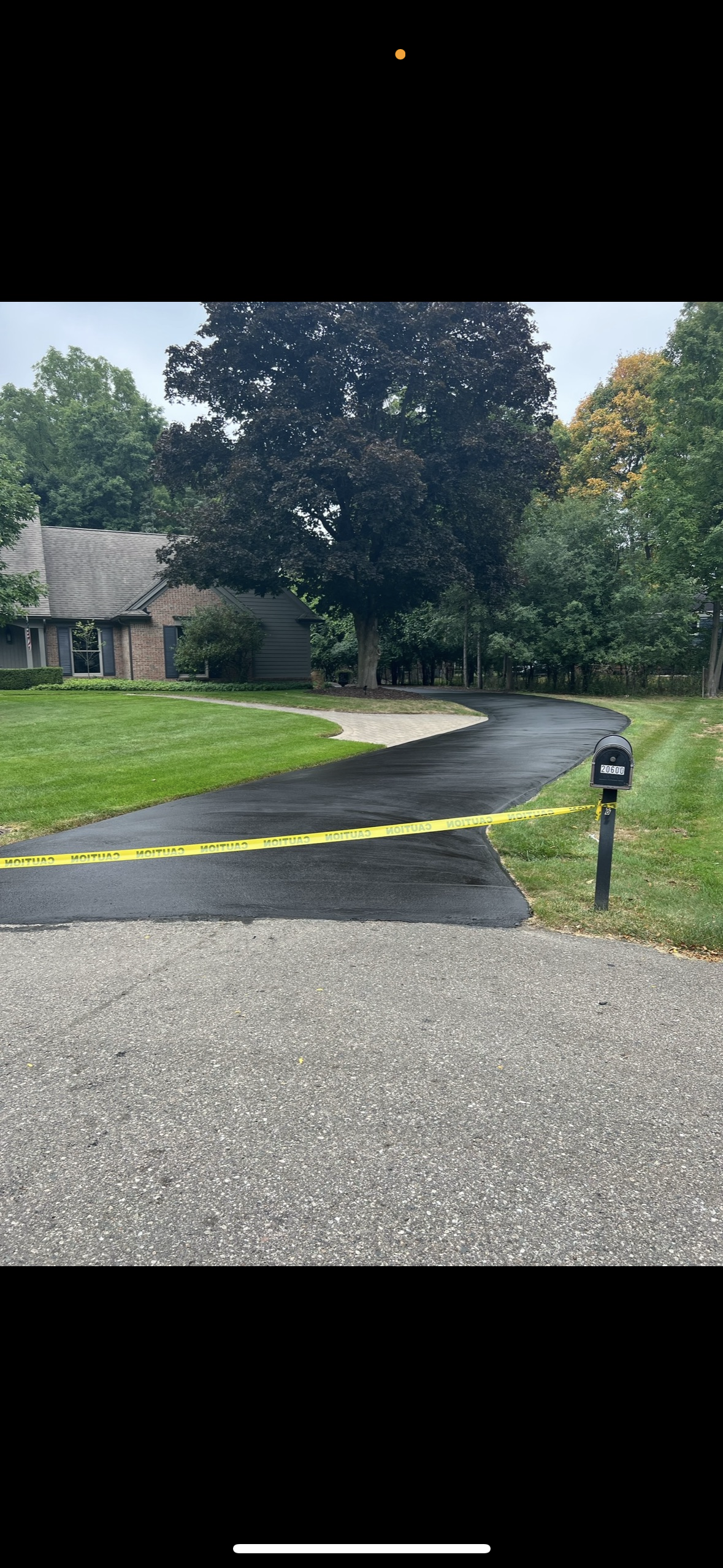 Driveway with freshly paved black asphalt, blocked off by yellow caution tape, leading to a house with a large tree in the front yard, a mailbox near the curb, and a grassy lawn.
