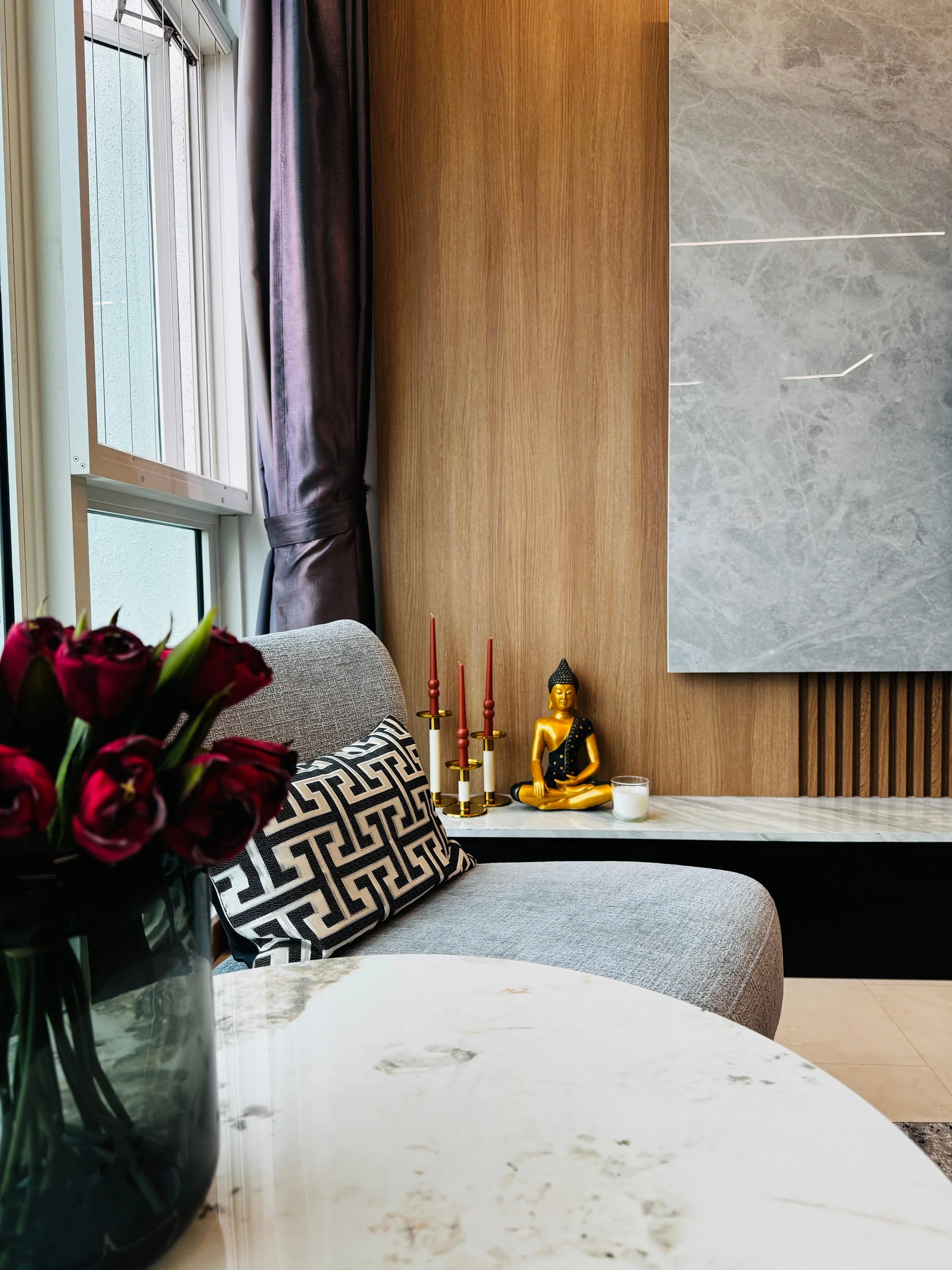 Interior of a modern living room with a gray sofa, black and white patterned pillow, vase of red flowers on a white marble table, window with dark curtains, wooden paneling, and a Buddha statue with candles and a candle on a shelf.
