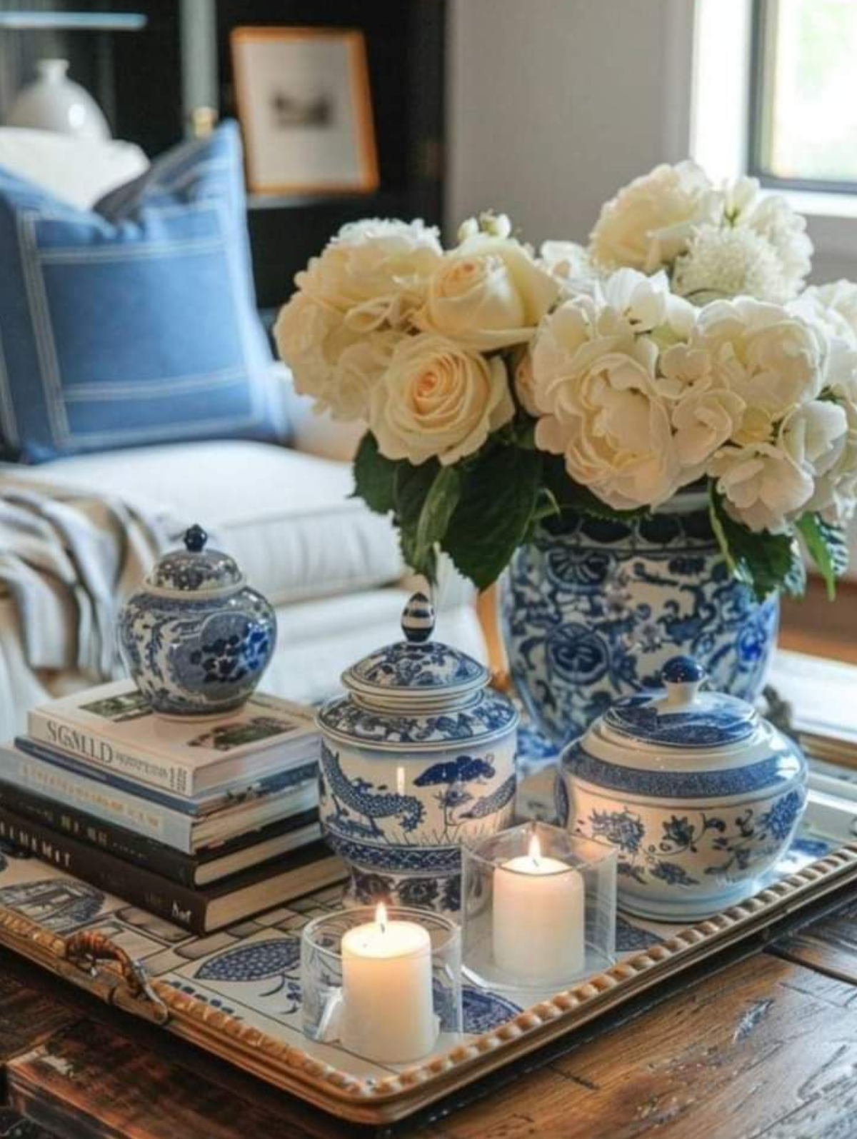 A decorative tray with stacked books, blue and white porcelain jars, a large blue and white floral-patterned vase with white flowers, and two lit candles in clear holders, set on a wooden table.