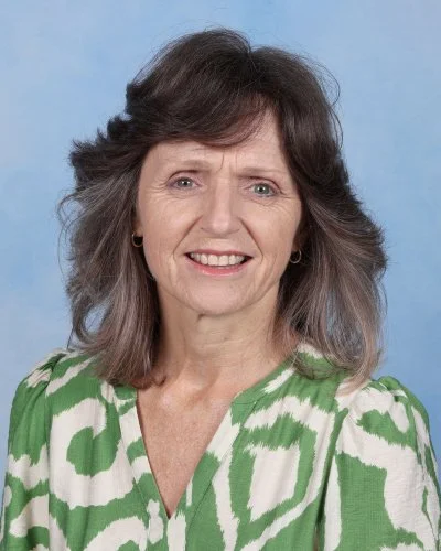 A middle-aged woman with shoulder-length brown hair and a floral-patterned blouse, smiling. Background is light blue.