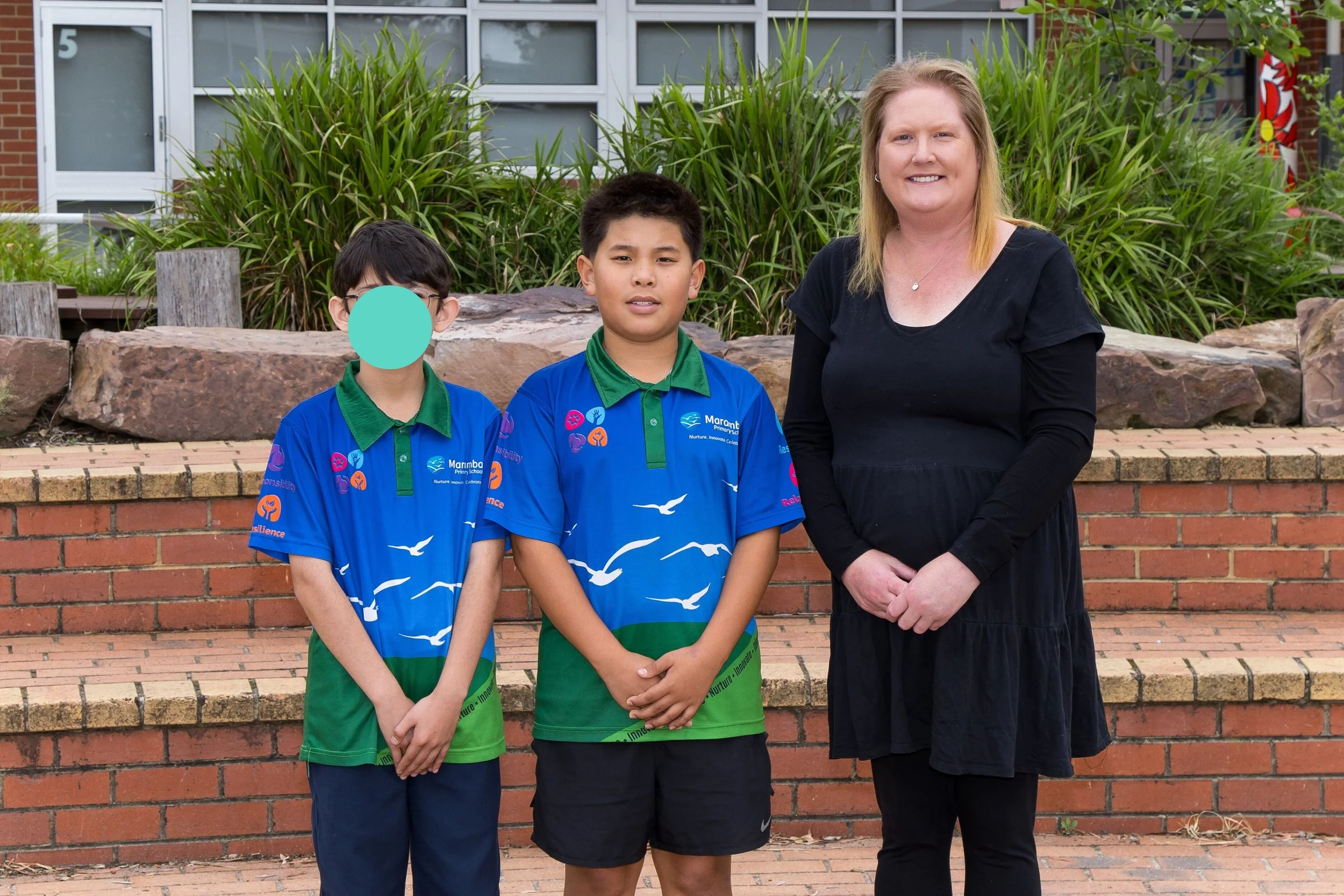 Two boys in blue shirts with a colorful snake design stand outdoors in front of a mural and greenery.