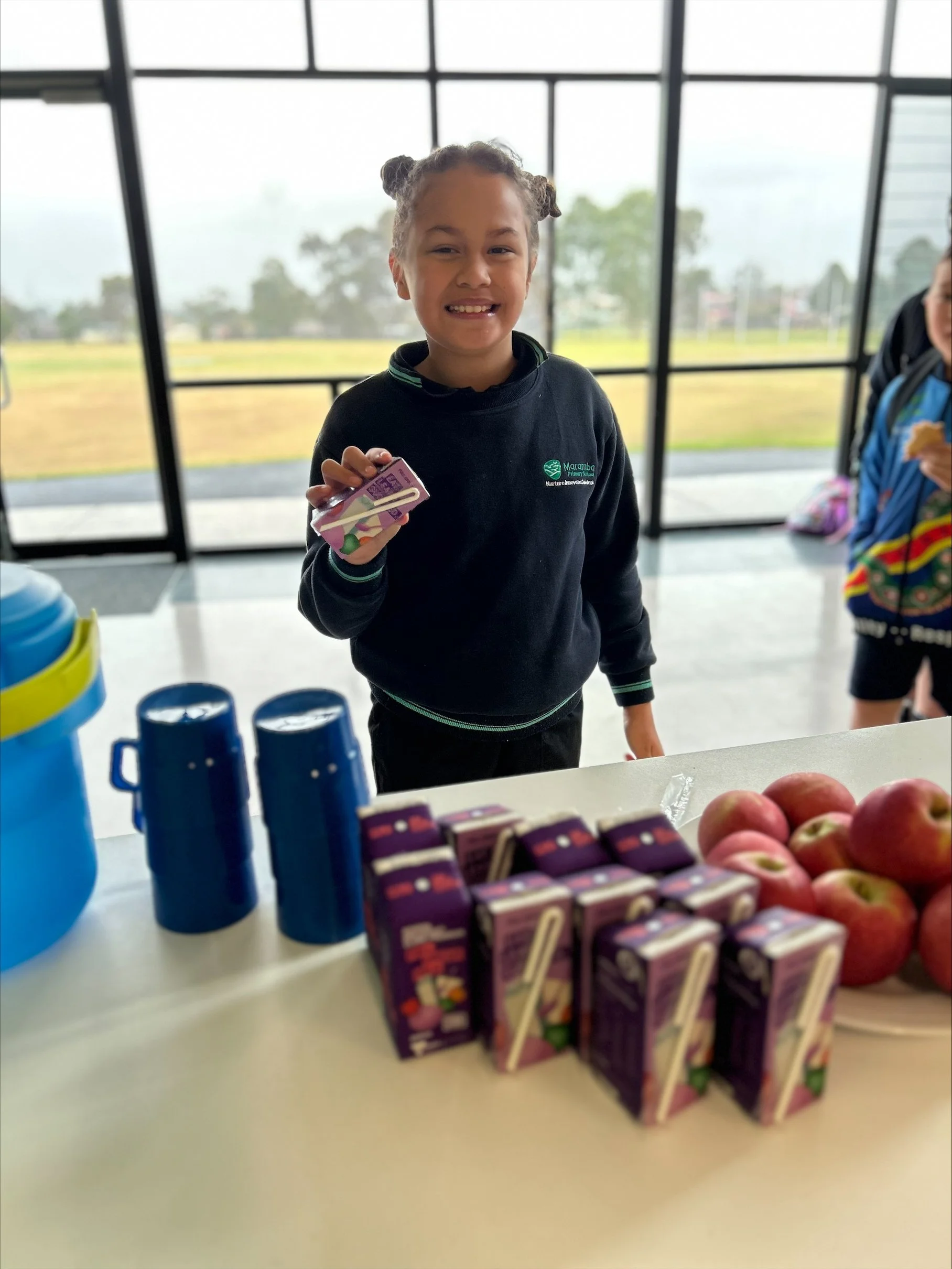 A young girl smiling and holding a small juice box at a table with more juice boxes and apples, inside a building with large windows showing an outdoor grassy area.