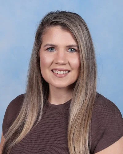 A woman with long brown hair smiling, wearing a pink floral top, against a blue background.