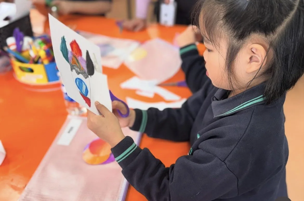 A young girl with short black hair in a black school uniform looks at an abstract painting on paper, sitting at an orange table with art supplies around her.