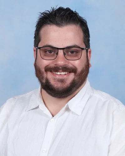 A man with dark hair and a beard smiling, wearing a light striped button-up shirt, against a light blue background.