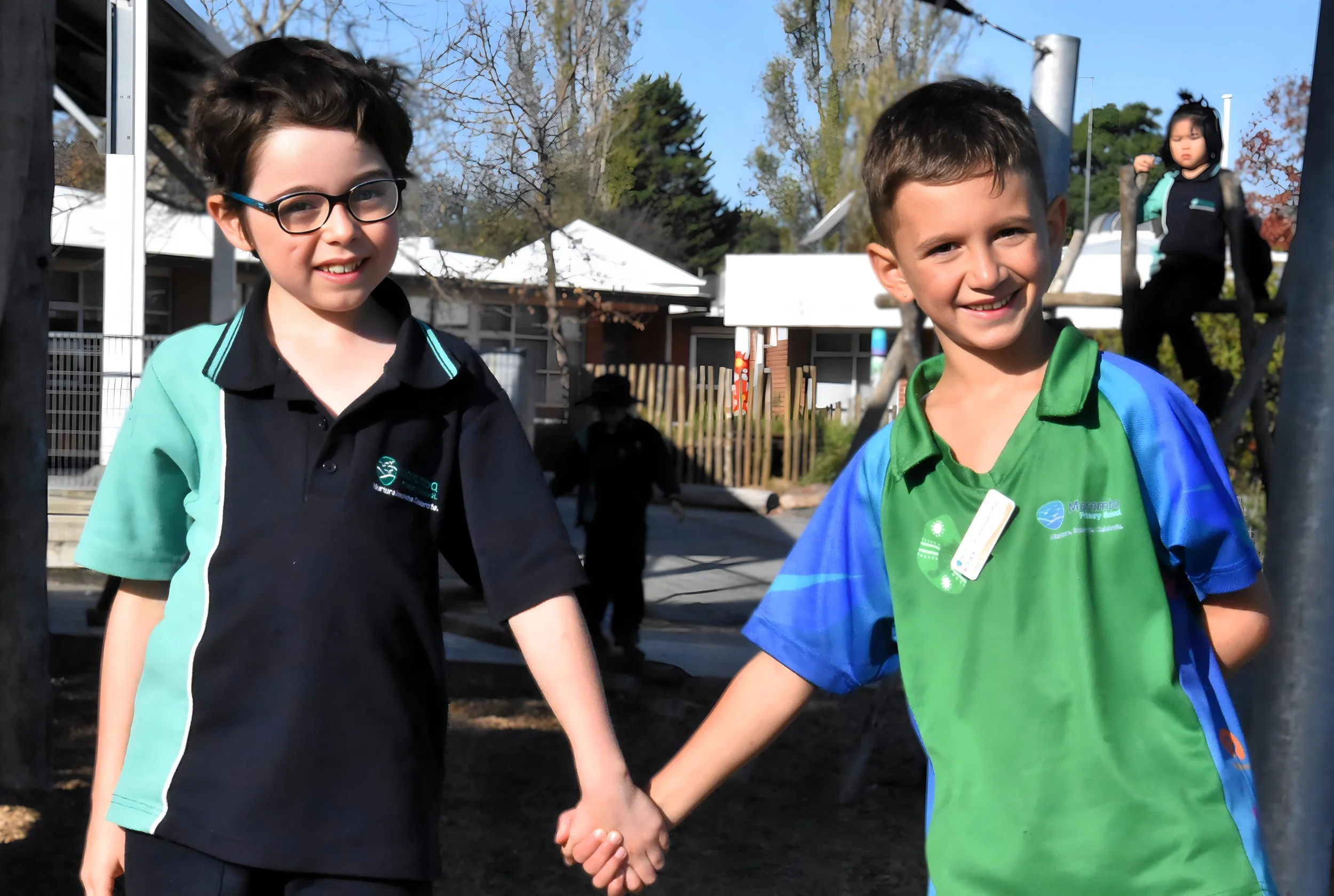 Two young boys holding hands and smiling outdoors on a sunny day, with one girl in the background and trees and buildings behind them.