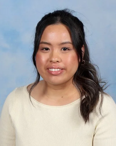 A woman with long wavy dark hair wearing a white blouse smiling against a light blue background.