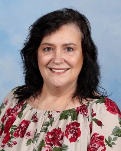 Smiling woman with dark hair, red glasses, wearing a colorful floral top, against a blue background.