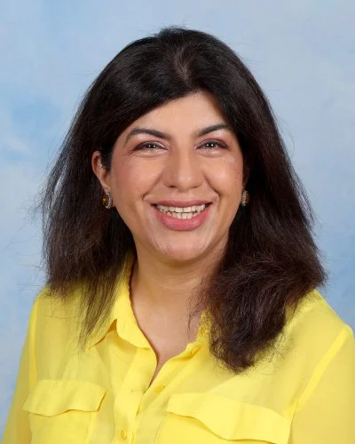 Woman with dark brown hair and light skin, wearing a patterned blouse and name tag, smiling at camera against a gray background.